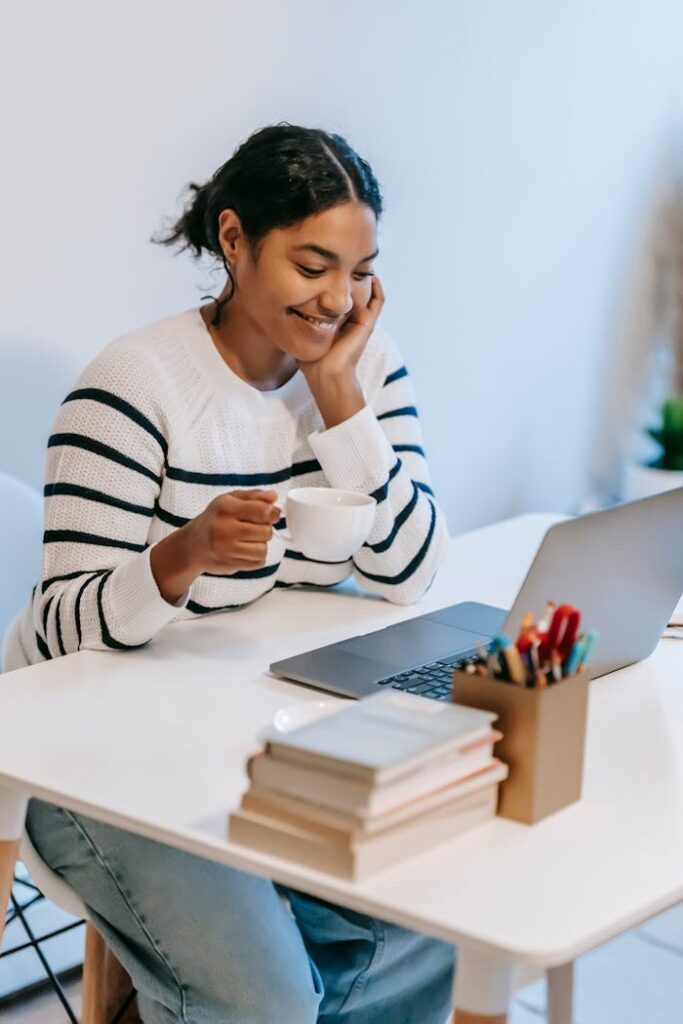 pexels-photo-6393342 Positive young African American female freelancer in casual clothes talking on smartphone and drinking coffee while working remotely on laptop at home